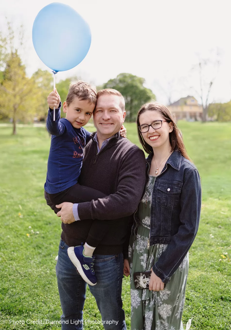 Josh Morgan and his two kids standing on a green lawn. He is holding his son with a blue balloon, while his smiling daughter stands beside them. The background is a bright park.