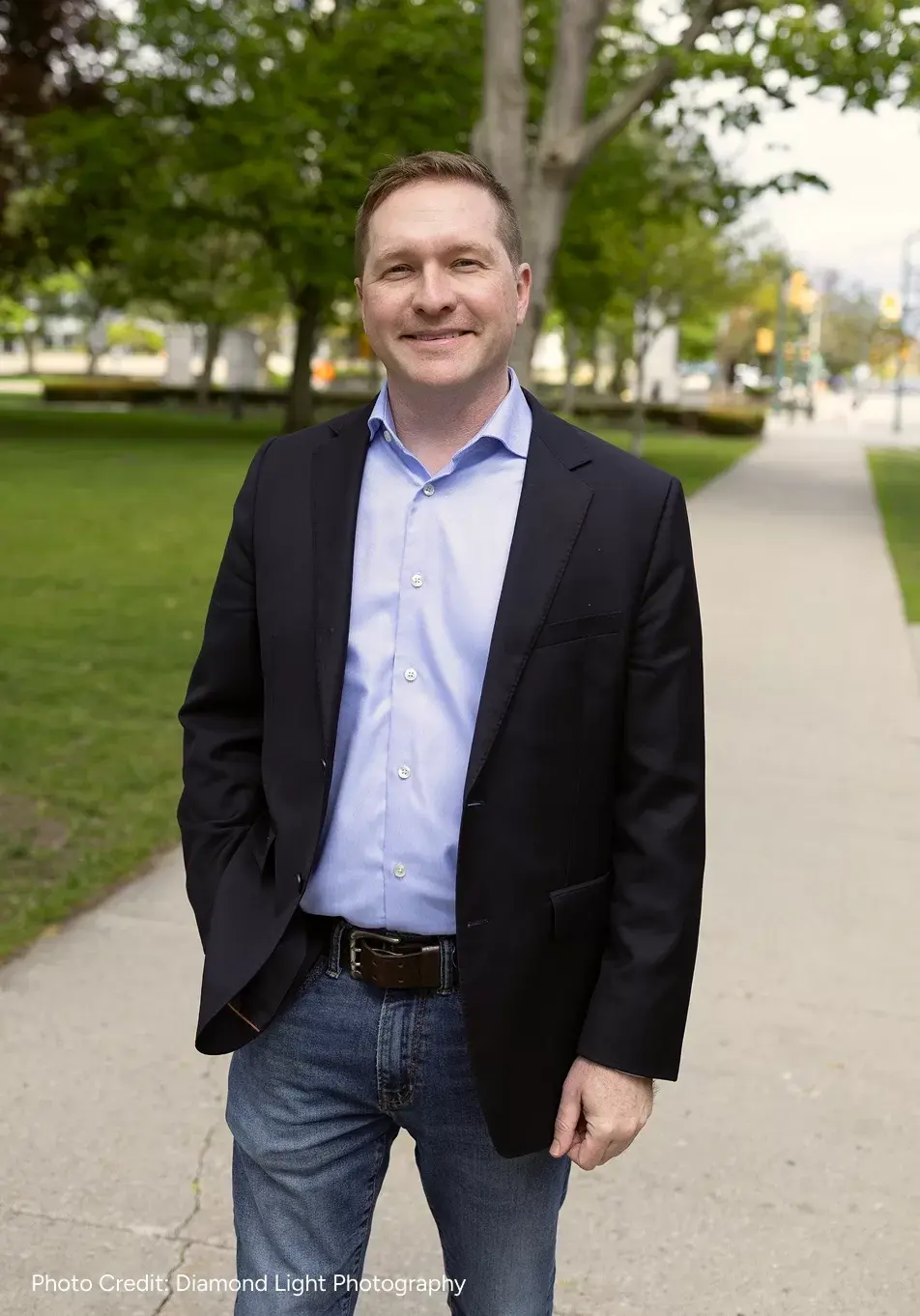 Josh Morgan in a blue shirt and black blazer smiles while standing on a park path with lush green trees and grass in the background, conveying a friendly tone. Photo Credit: Diamond Light Photography